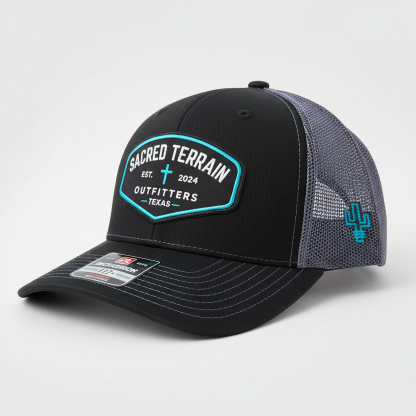 Black and gray cap with 'Sacred Terrain Outfitters' logo on a rock with grass background